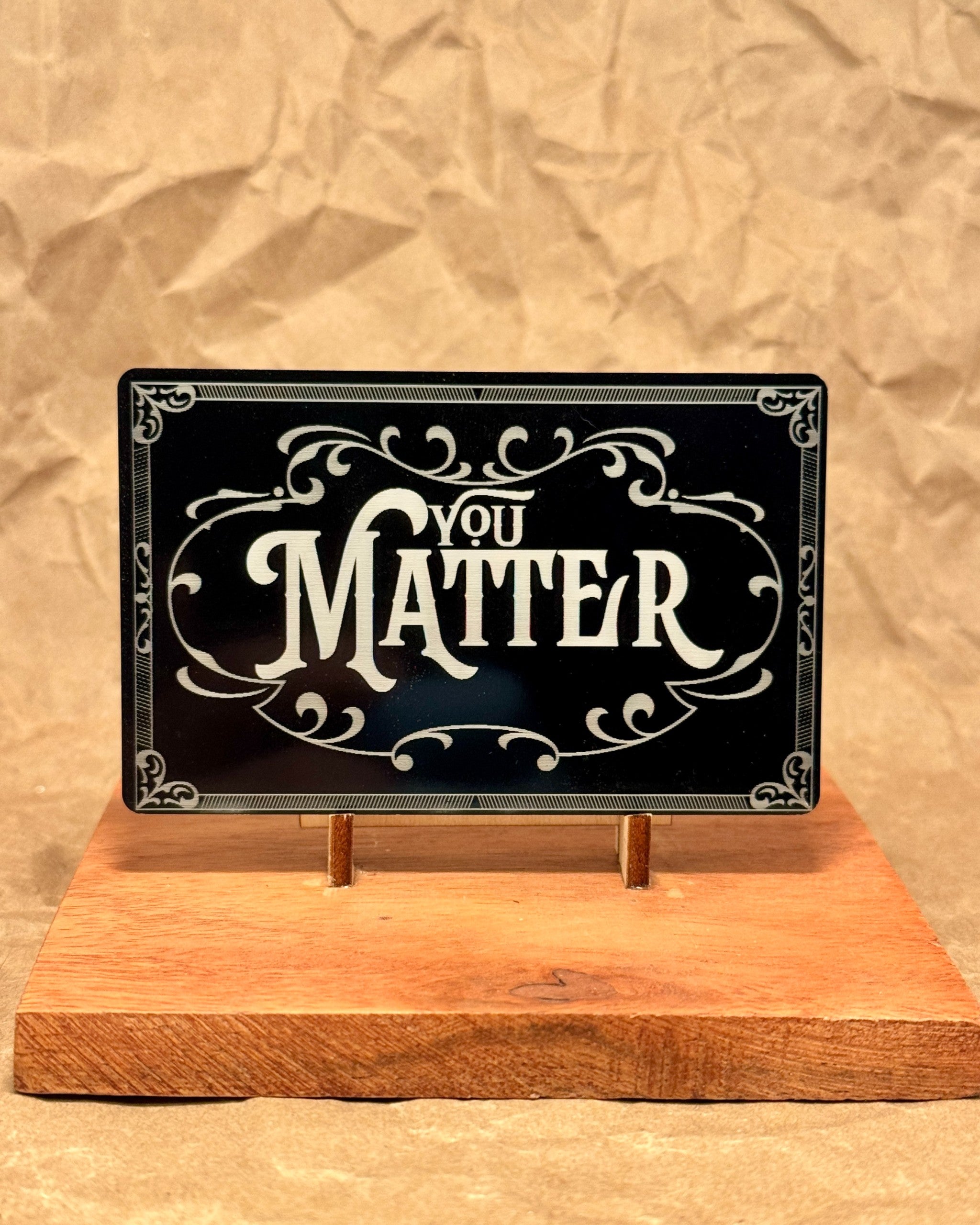 Black You Matter metal wallet card with an ornate engraved border, displayed on a wooden stand
